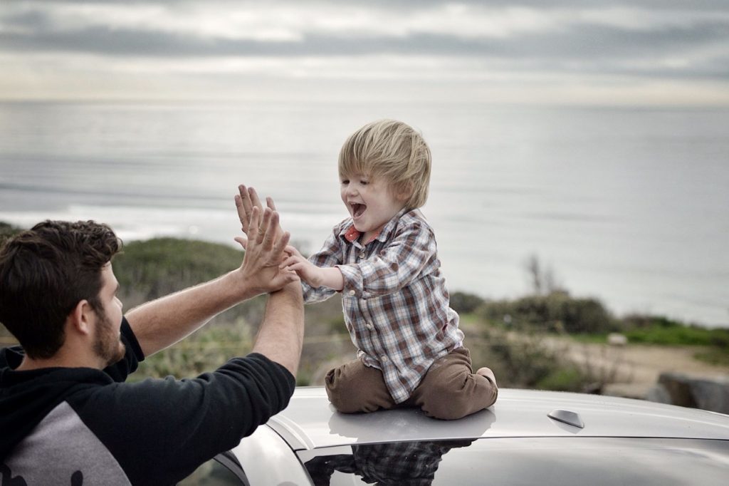 ID: a young boy sits on top of a car. He is clapping both hands with a man.