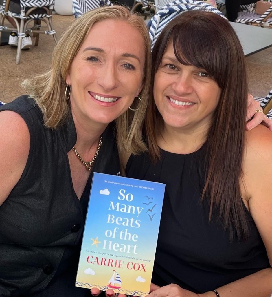 ID: author Carrie Cox with her arm around her friend Maria, who is holding a copy of So Many Beats of the Heart