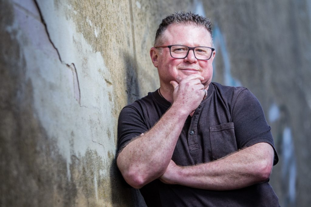Image of author Josh Langley leaning against a wall. He is smiling and resting his chin on his hand.