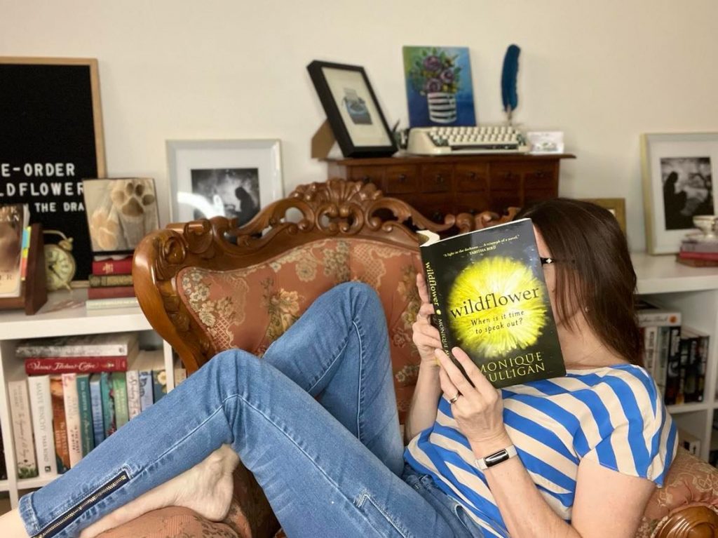 ID: author Monique Mulligan is sitting sideways across a high-backed antique leather chair, with her legs hanging over the arm rest. She is reading a copy of Wildflower with her face obscured by the books.