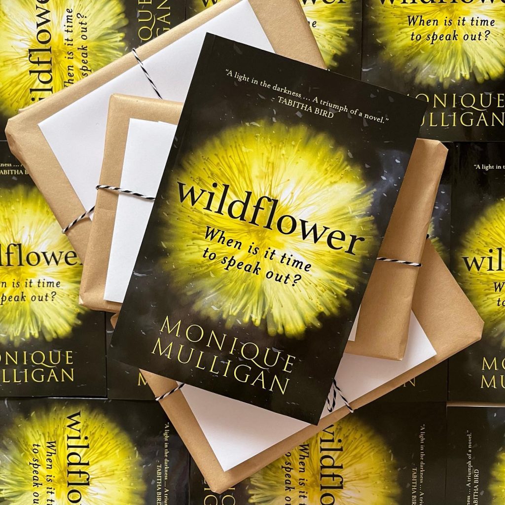 ID: a copy of Wildflower lies on top of several packages in brown paper and tied up with string. These sit on a large pile of Wildflower copies