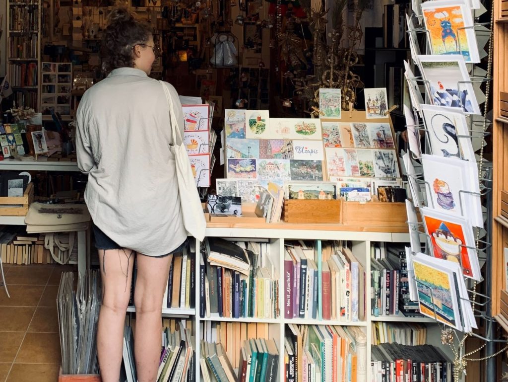 Girl in cut off shorts and a calico bag over her shoulder standing at the entrance to a bookshop.