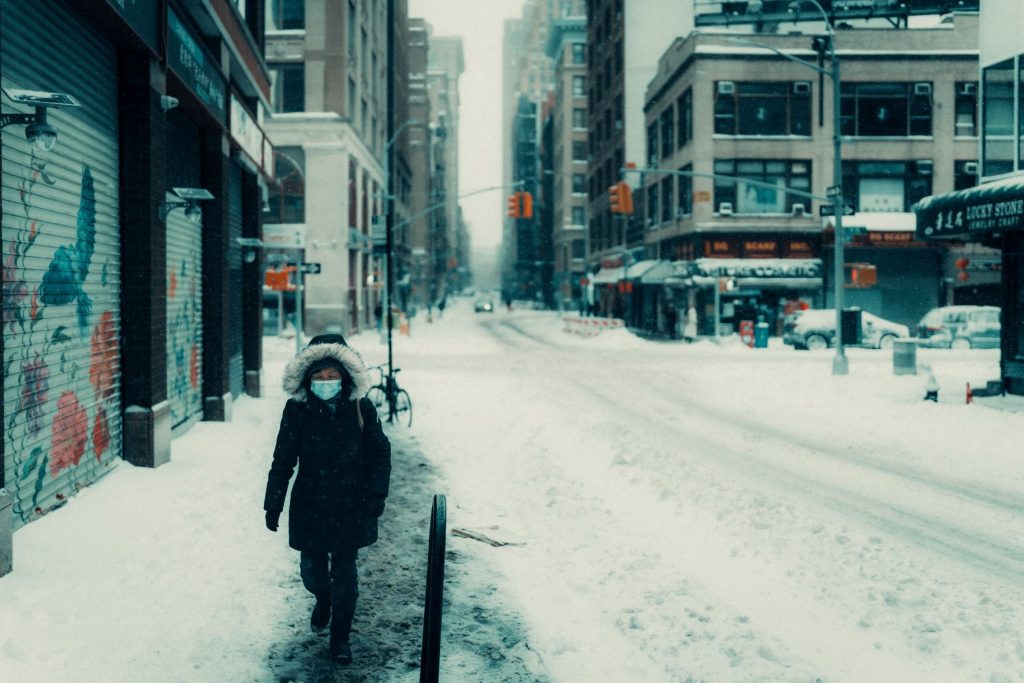 ID: an icy streetscape with tall buildings on either side of the street. The street is covered in snow and a lone person on the side walk walks towards the camera and is wearing a hooded jacket and a face mask.