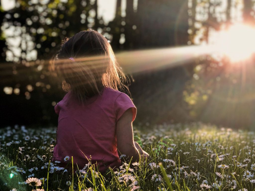 ID: a young girl is sitting in a field of flowers. The sun is streaming through the trees and the girl's face is tilted towards the sunlight.