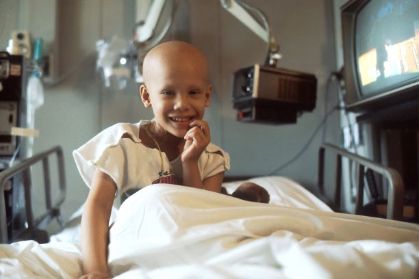 Image is of a child with a bald head sitting up in a hospital bed and smiling at the camera